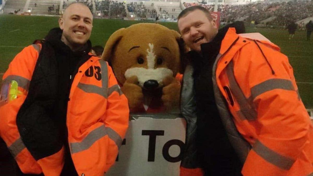 Ben Moss and Tom Doolan are smiling while wearing hi-vis orange jackets, with a football mascot that looks like a dog standing between them, at a football ground