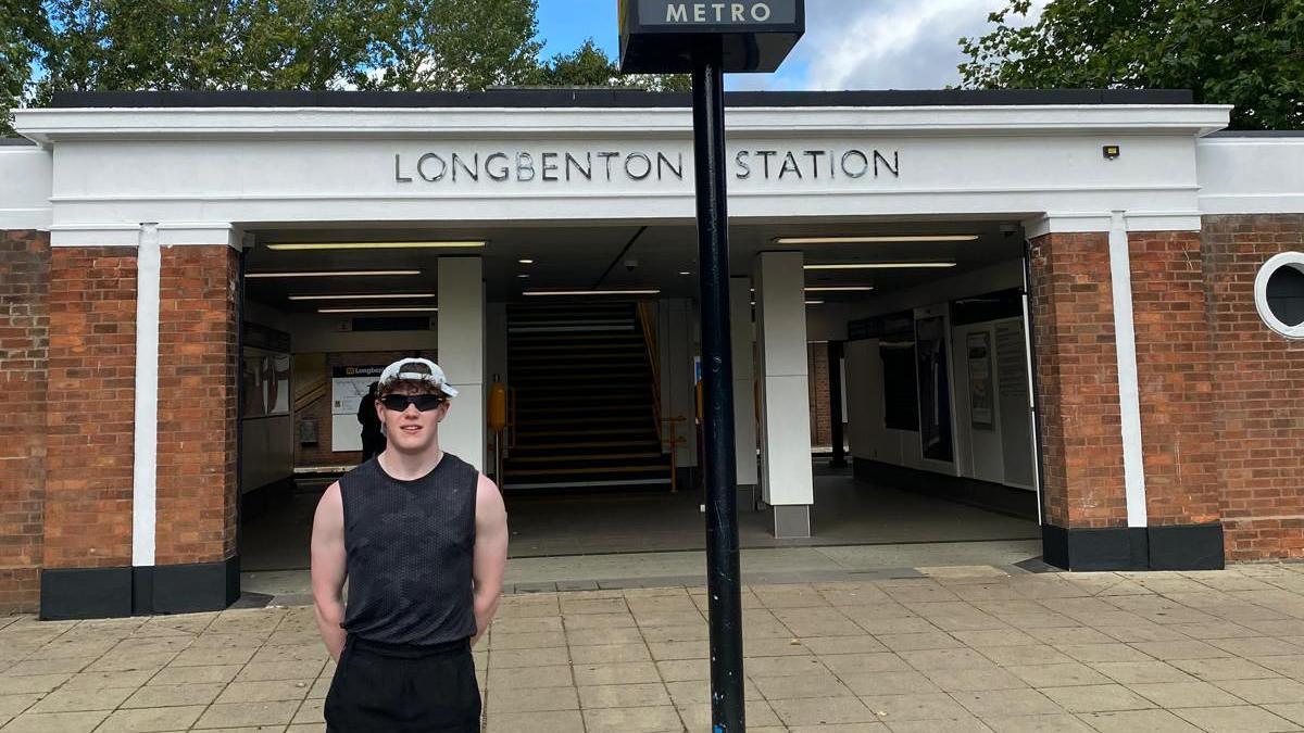 Charles McManus standing outside Longbenton Station wearing a black vest and a cap and sunglasses. 