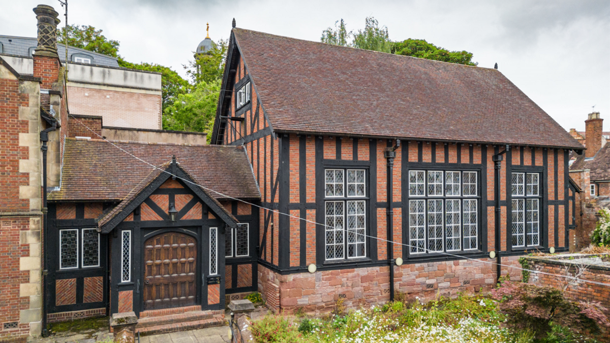 The exterior of a red brick building with black wooden beams, large windows and a brown tiled roof. There is a covered entrance with a large wooden door on the left hand side