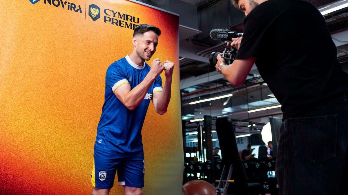 Kyle McCarthy in a blue Haverfordwest shirt poses in front of a camerman with the Cymru Premier backdrop behind him