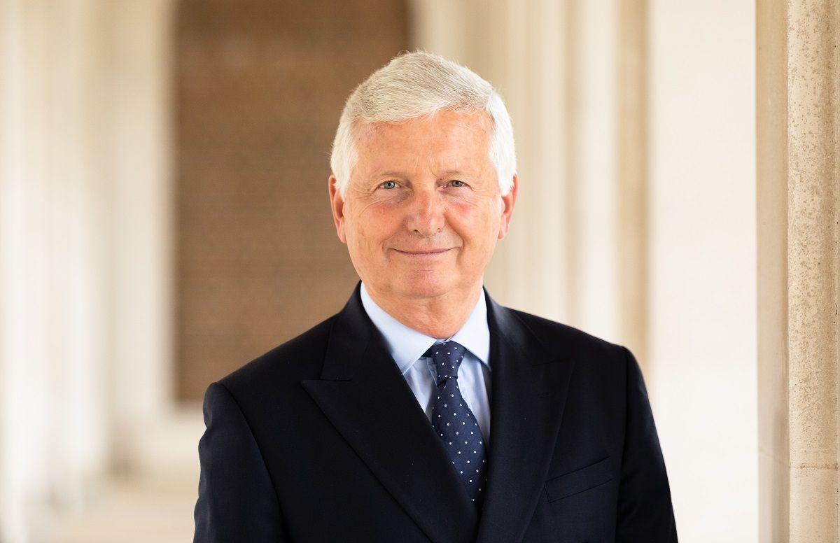 A picture of a man with short white hair, stood in a corridor outside a building. He is wearing a black suit jacket with a blue tie with white spots on it and a light blue shirt.