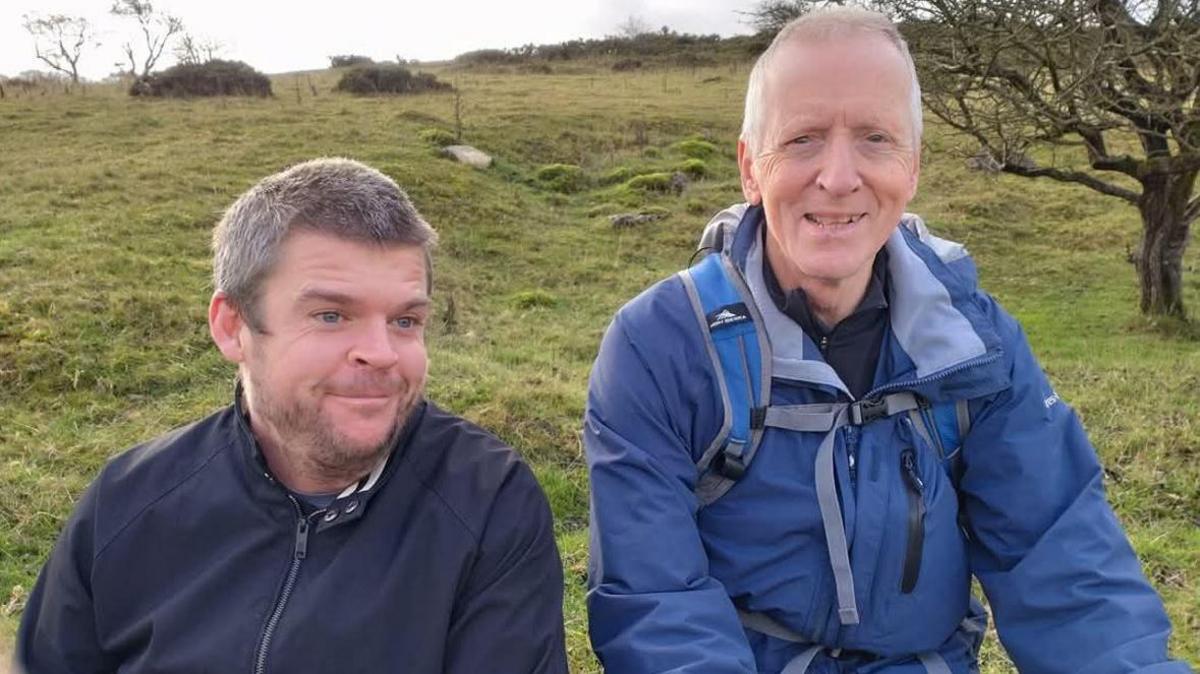 Two men in a field wearing blue tops. One man has dark hair and the other has grey hair