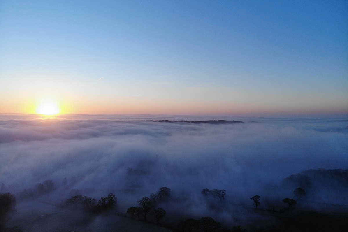 Thick clouds of mist coat a countryside with the rising sun visible in the distance at the horizon. The odd clump of trees show through, black against the white mist.