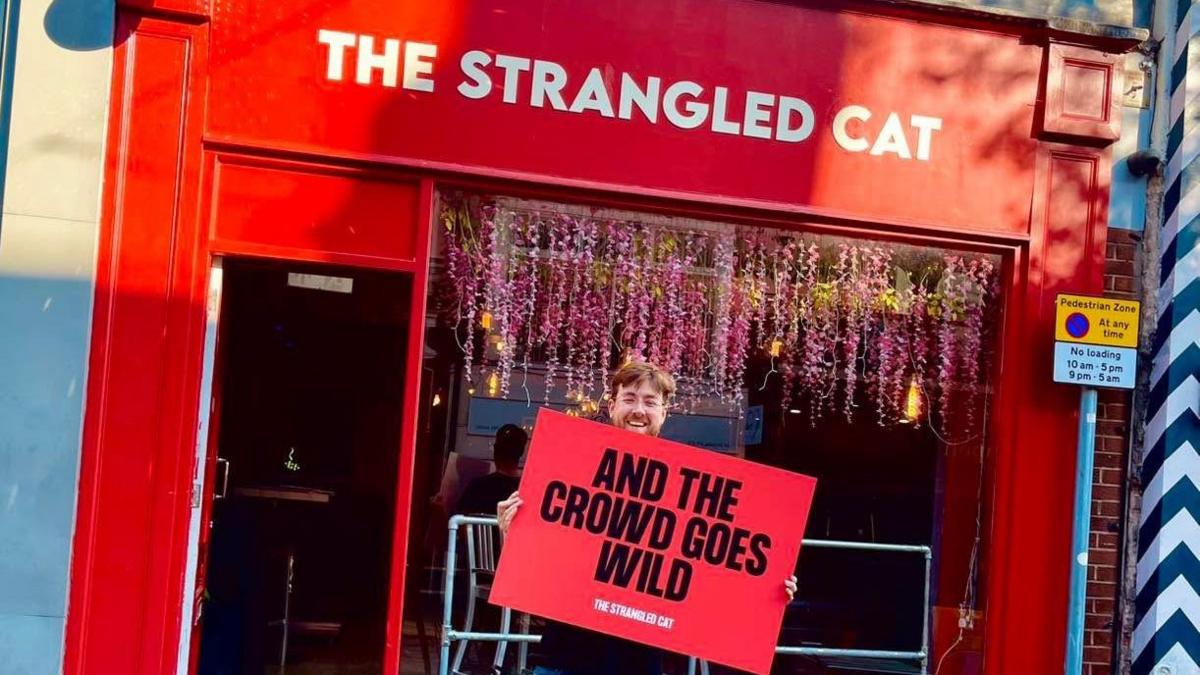 A young man with short hair and a beard and wearing a black T-shirt is standing outside a bar with a red frame with the words in white lettering above the large window, "The Strangled Cat". The man is holding a sign that reads: "And the crowd goes wild".
