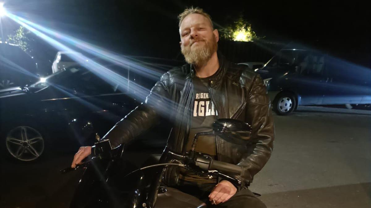 Paul Hart sits on a motorbike at night. He is wearing a black top and leathers and a bright light is shining down. There are cars parked nearby.