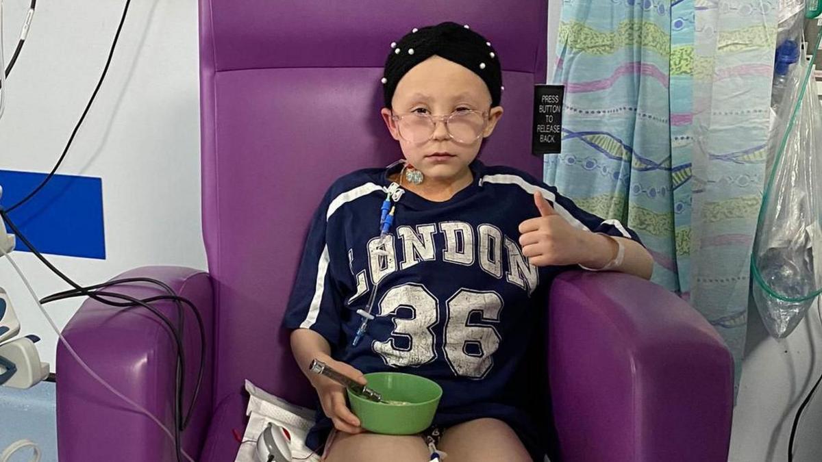 Mae merch 10 oed yn eistedd mewn cadair ysbyty borffor. Mae hi'n gwisgo siorts du a chrys-t nefi. Mae hi'n gwisgo sgarff pen du gyda smotiau gwyn arni a sbectol.