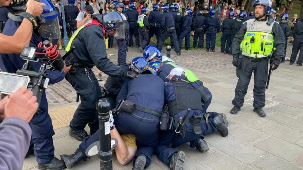 Four police officers kneel over a man on the floor while around 20 others can be seen controlling crowds in the background