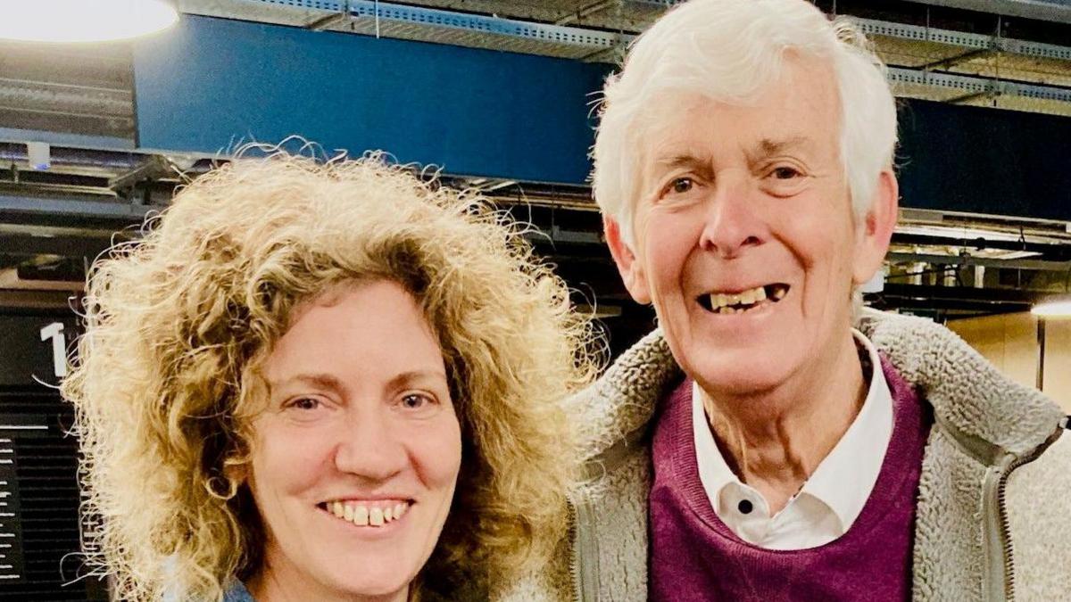 Lynda and Terry Walton smiling whilst looking at the camera in BBC Wales' building in Cardiff. Lynda, on the left, has curly blonde hair. Terry, on the right, has short white hair and is wearing a white shirt, burgundy jumper and a grey fleecy jacket.