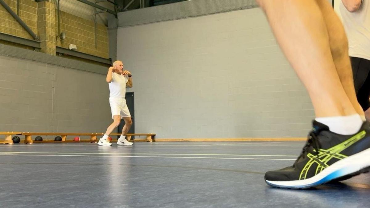 A man is stood in a gym jumping up and down. He is in all-white and there are black shoes in the foreground.