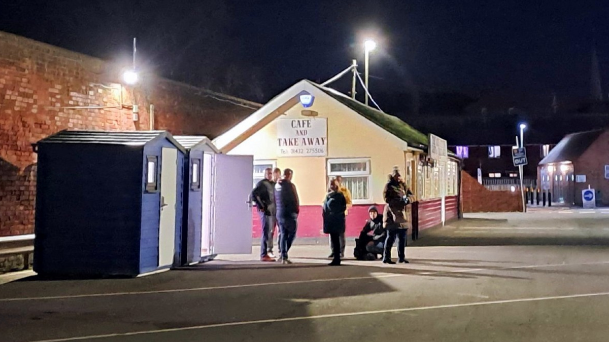 Two garden shed-type structures, painted blue and each with a white door and a small window, stand on the edge of a car park at night. Several people are standing nearby and there is a cafe close to the sheds.