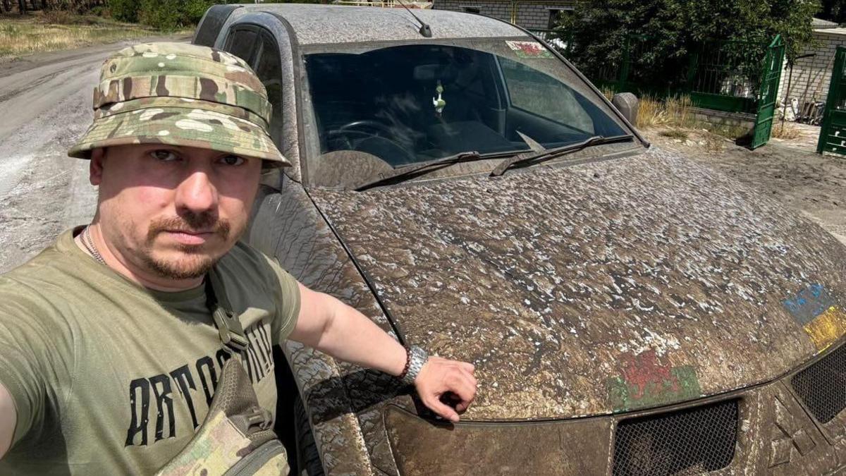 A Ukrainian soldier in a camouflage bucket hat stood in front of a very muddy Mitsubishi SUV. There are Wales flags and Ukraine flags on the front of the vehicle under the dirt