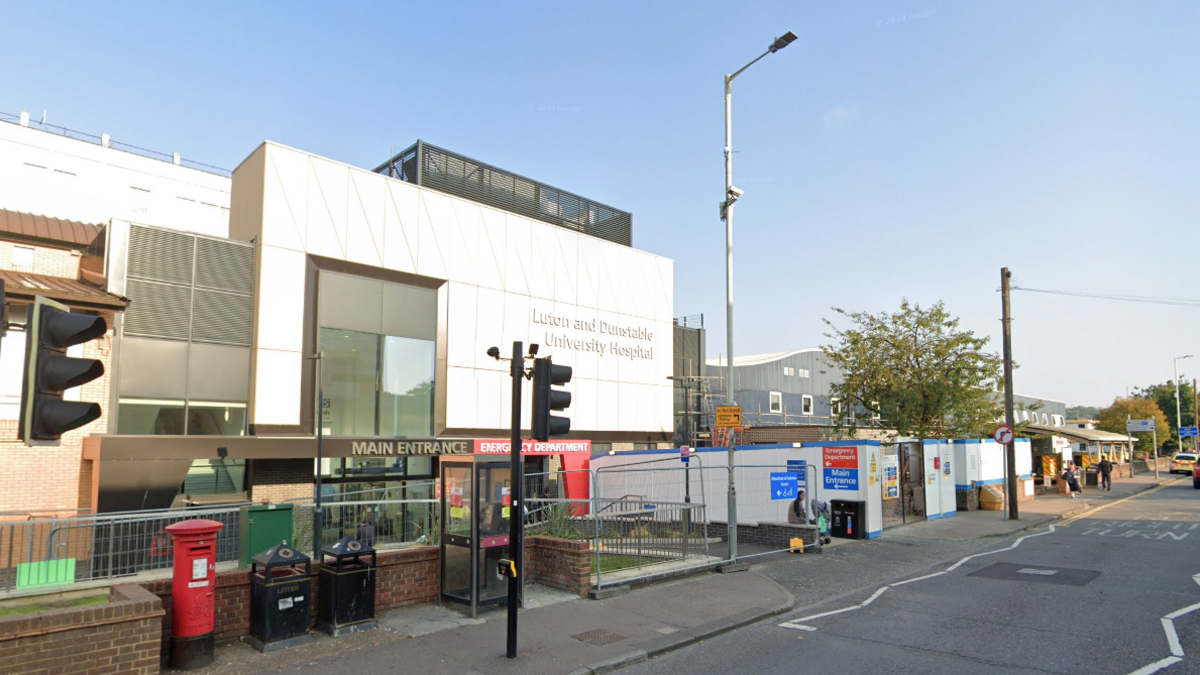 The outside of Luton and Dunstable Hospital showing the main entrance, railings, boardings, a phone box, pillar box and litter bins. The building has a rectangular white frontage.