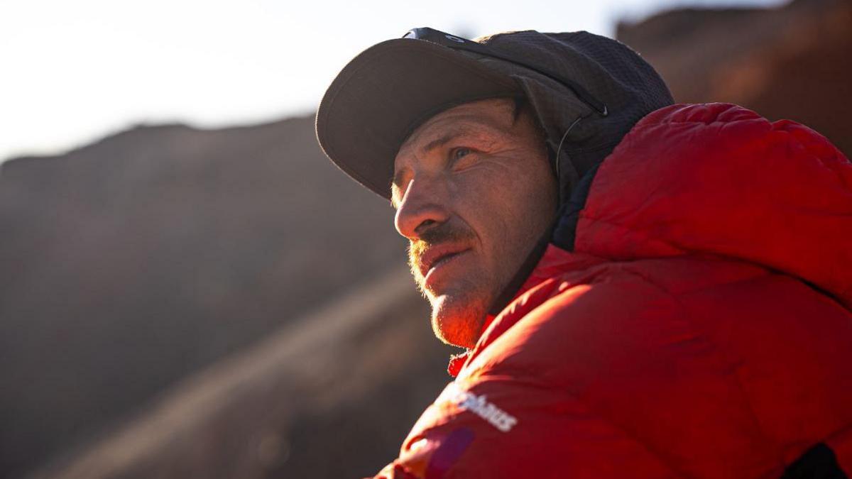 A close up image of Ed Jackson wearing a black cap and an orange thermal jacket. He is looking off to the left of the camera with a thoughtful expression. The light is soft and it appears to be approaching dusk.