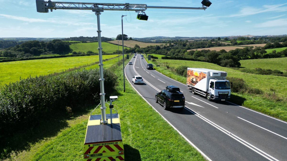 A view of the road from an elevated angle showing it flowing off into Devon countryside. There are fields and trees. There are multiple vehicles using the road including cars and lorries. In the foreground is the AI camera installed on the top of a vehicle. It resembles a small crane attached to the top of the vehicle with cameras attached to it. 