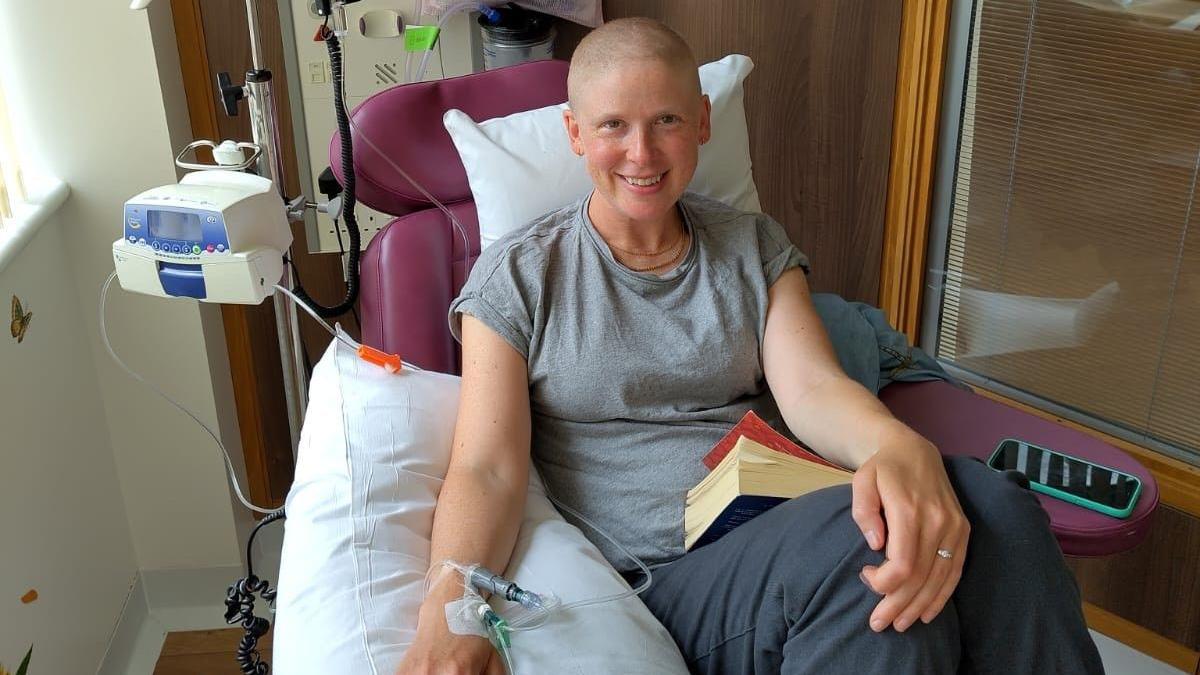 Phillippa Hentsch sitting in a chair with a cannula in her wrist. She is holding a book.