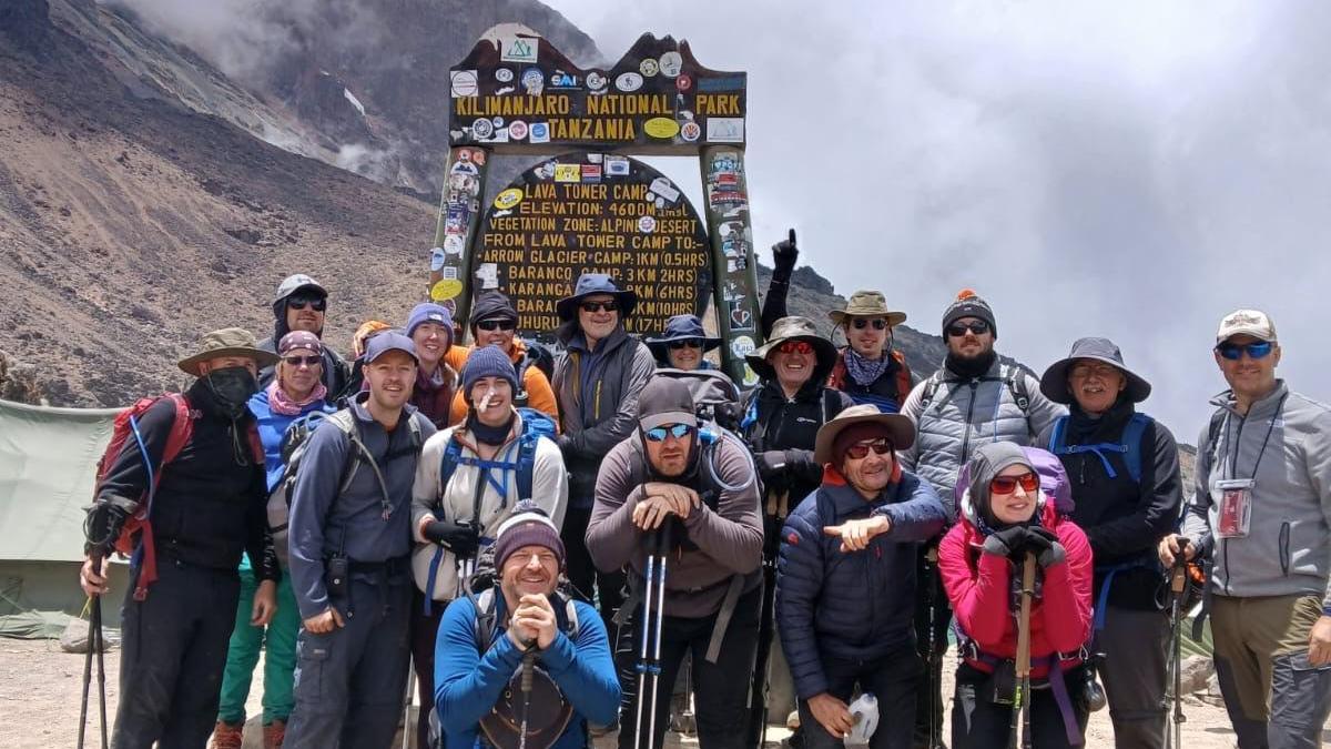 A group of people standing in front of a sign at one of Mount Kilimanjaro's camps. The mountain is in the background