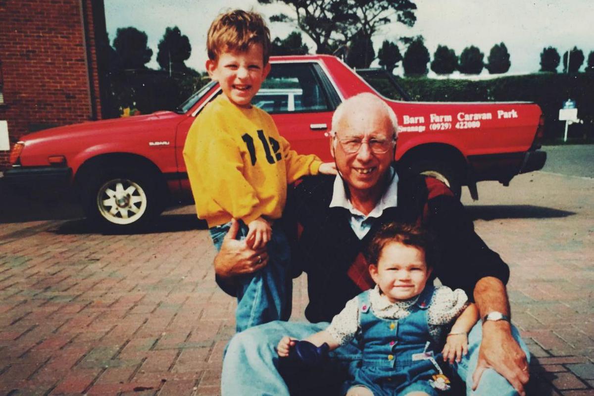 An old photograph of Issy Sedgwick as a toddler with her grandfather and brother. The three of them are posing for the camera on a block-paved driveway with a red Subaru pick up truck behind them. Issy is sitting between her grandfather's legs while her brother, who is wearing a bright yellow jumper, is standing next to him with his hand on his grandfather's shoulder.