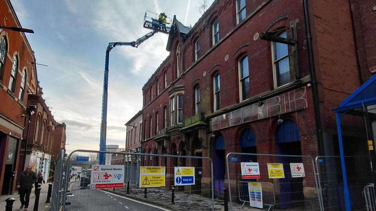 A tall red brick building can be seen but in front are metal fences with warning signs all over reading 'danger'. A crane can be seen as workers in hi-vis jackets.
