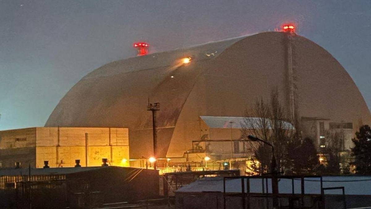 The large curved metal shield over reactor four, pictured in low light, with flame emerging from a hole in one side where it was hit by a drone on 14 February 2025. There are red lights on top of the dome, and there is yellow lighting around the lower buildings close to it.
