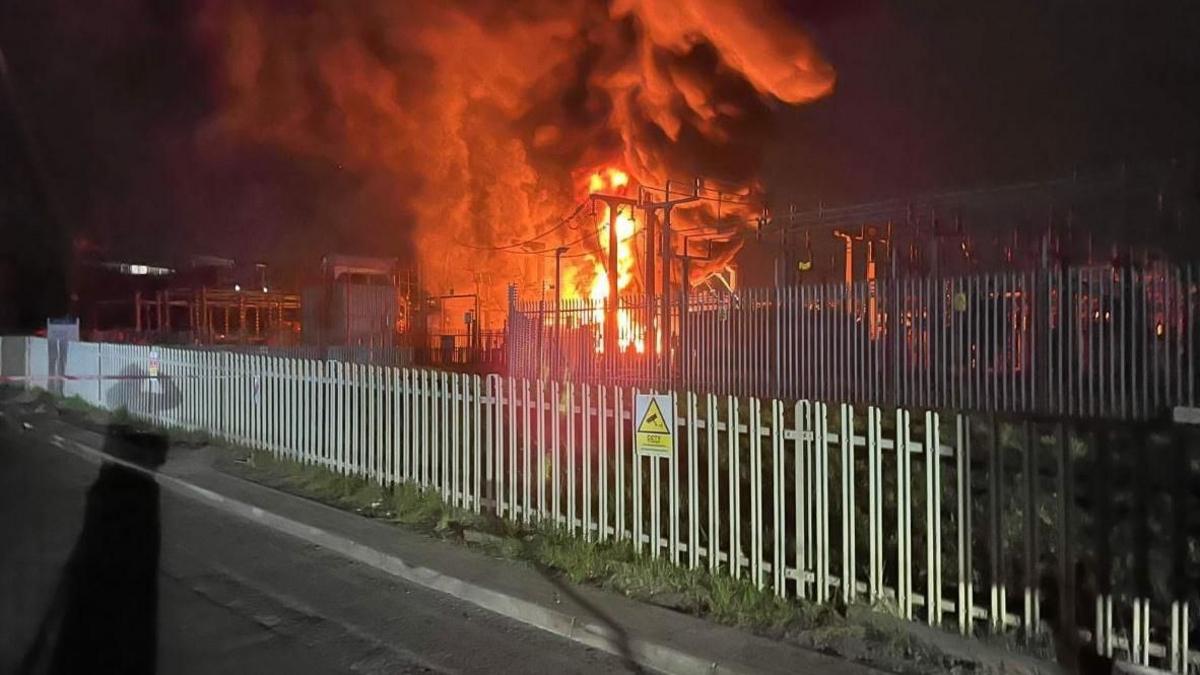 Handout photo supplied by London Fire Brigade showing a fire at Hayes electrical substation. Orange flames and smoke billow into the sky behind a grey fence.