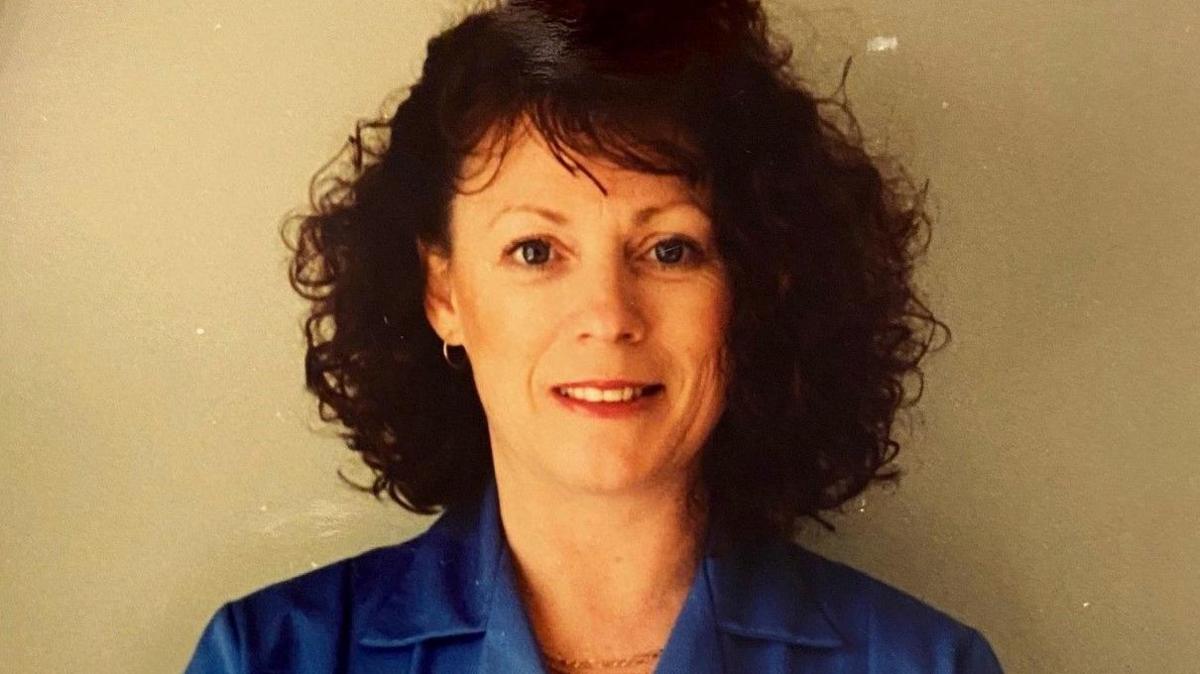 Beverley Pedley in a blue nurse uniform is smiling at the camera. She has dark brown, curly hair.