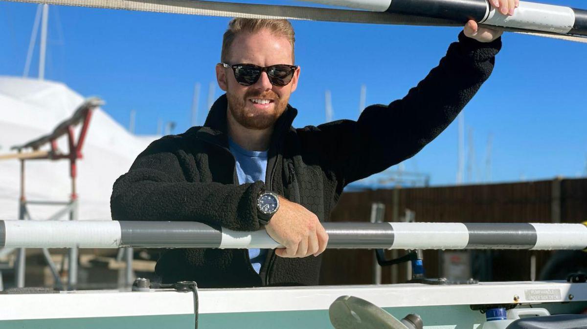 Kyle Meredith is leaning against a boat. He wears a black fleece, black sunglasses and a black watch