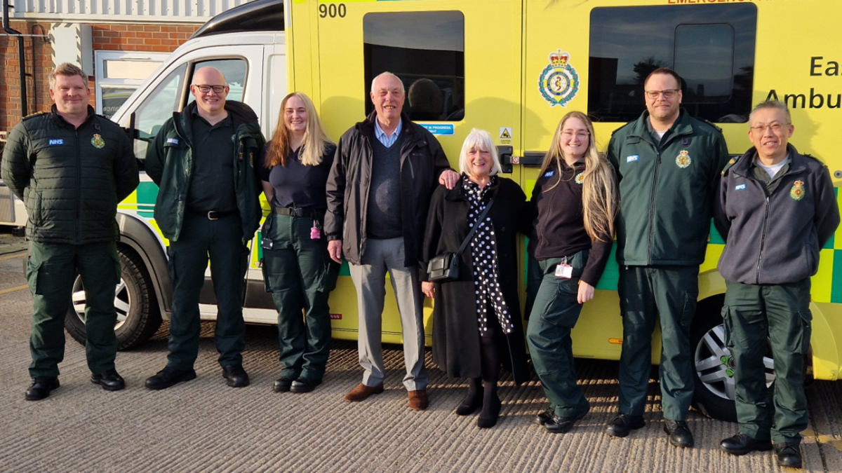 Eight people smiling, standing in line, facing the camera, with an ambulance behind them. A man and woman with grey hair are in the middle, and are flanked by two women and four men, who are all in ambulance service uniform. 