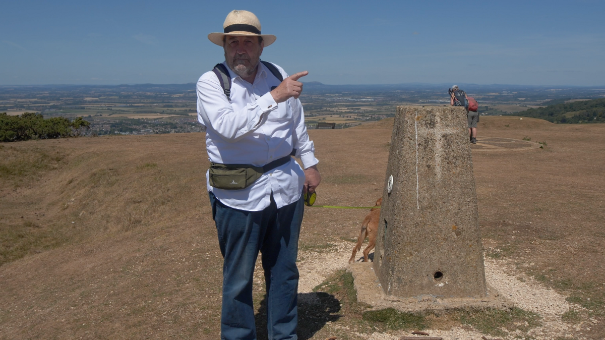 Robin Shuckburgh next to a monument on Cleeve Hill on the Cotswold Way National Trail. He is pointing at something off camera. He is holding his dog on a leash. Another man can be see in the distance. It is a clear day.