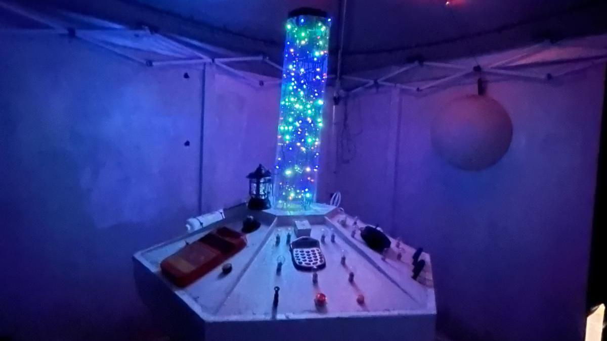 A model of a white control panel with various buttons, with a tube of sparkling blue and green lights in the centre. There is dark blue panelling behind.