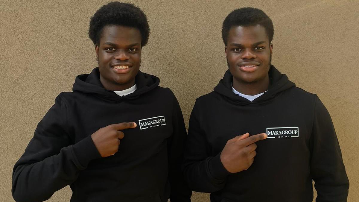 James and John Makanjuola are standing side-by-side, smiling and looking directly at the camera. They are wearing matching black hoodies and tracksuit trousers. The hoodies have a logo on the left which reads 'Makagroup' and the twins are each pointing towards the logo.