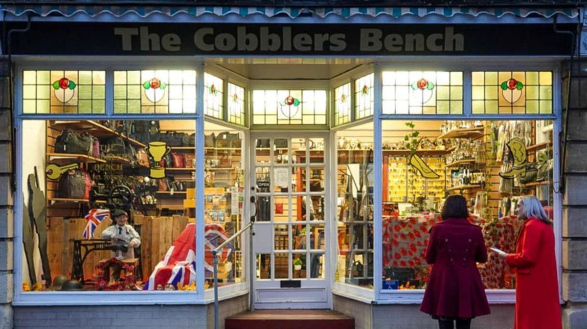 A shop as it starts to get dark, lights are on shining through the large windows and glass door. It is a traditional cobblers shop, with lots of poppies and union jack flags in the display. Two women in red coats are looking at it.