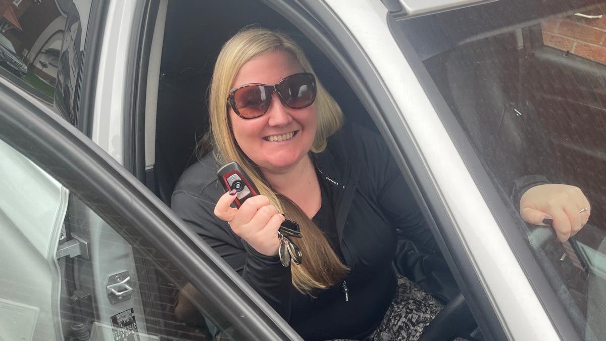 Hayley sits in her grey car, behind the steering wheel showing her car keys to the camera. Hayley is wearing brown sunglasses and smiling.