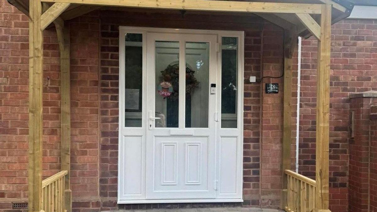 A white door sits beneath a timber porch at a house with a sign in the window that reads "We Are Open". 
