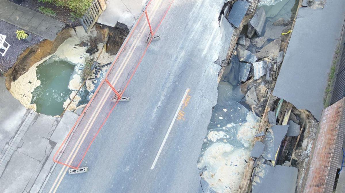 A drone shot looking down onto the two sinkholes in Godstone, both of which contain pools of water. A section of pavement and the garden of a property have collapsed into the smaller sinkhole on the left, while one lane of the road and part of the pavement have collapsed into the larger sinkhole on the right.