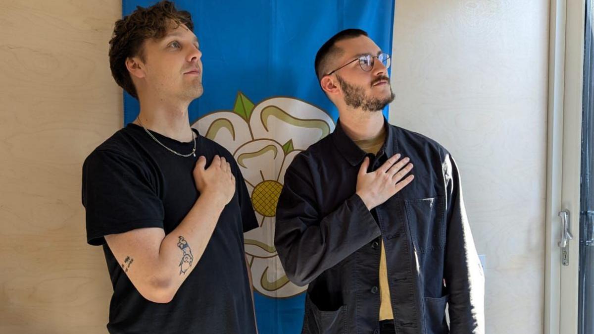 Two young men stand in front of a Yorkshire flag - a white rose on a blue background - hung on a wall. Each has his hand placed across his chest, looking off-camera at the sky. It's a tongue-in-cheek pose reminscent of football players lining up for their national anthem before a game.