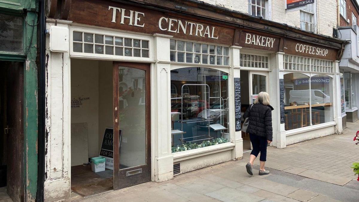 The outside of the bakery as a woman walks past. The bakery has several big windows with a sign overhead reading "The Central Bakery - Coffee Shop"