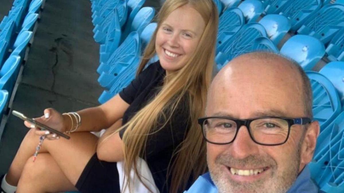 Paul Ross and his daughter Lydia Ross sit in football stands in a selfie style picture. Lydia has long blond hair and Paul wears black-rimmed glasses and is bald and has grey stubble. They are sitting in a football stand surrounded by empty sky blue seats.