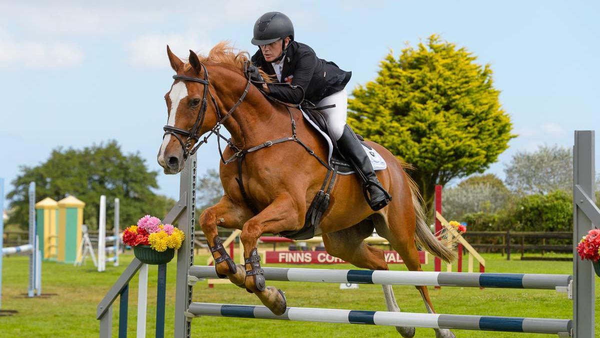 Justin Ogier and his brown horse jump over a grey, white and green fence during a showjumping competition. Justin is wearing a black jacket, white jodhpurs and black riding boots. He has a black helmet on. Other showjumping fences are dotted around the tree-lined field.