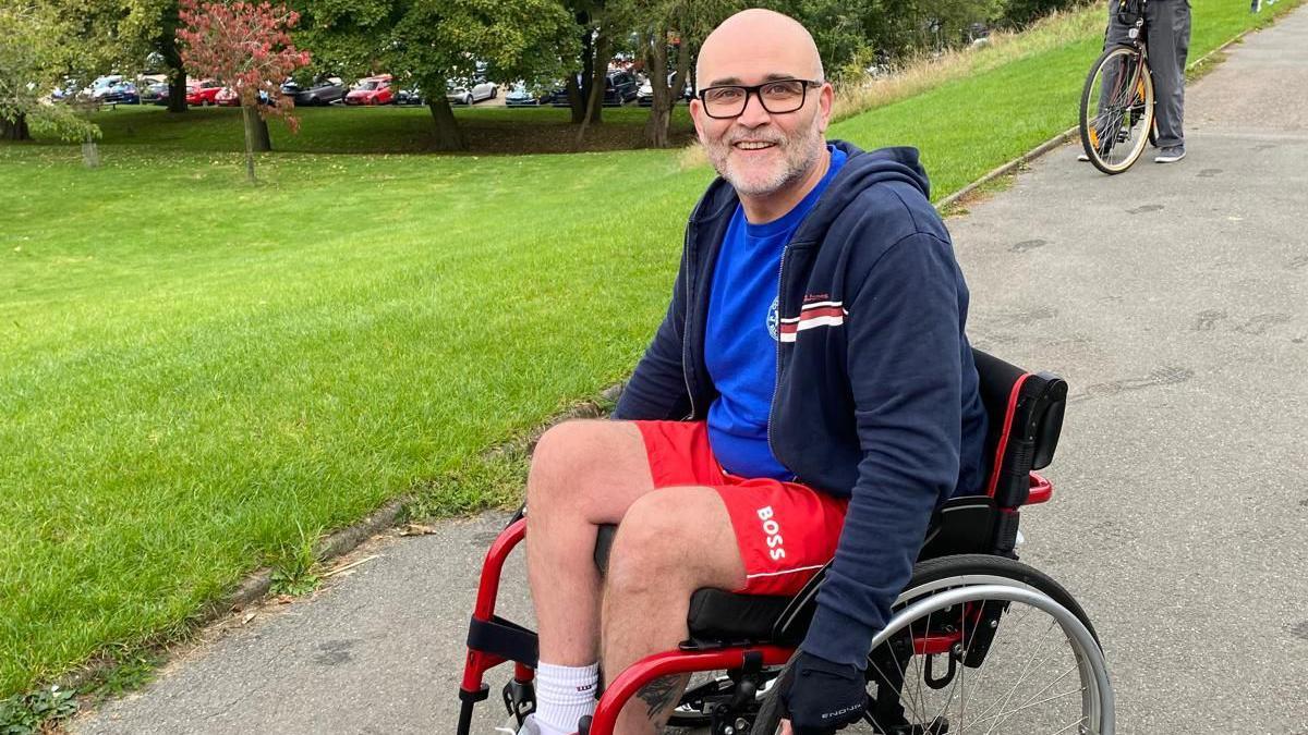 A man wearing shorts and a T-shirt while sitting in a wheelchair – he is smiling at the camera