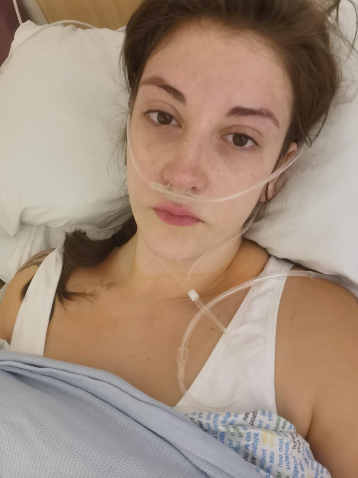 Woman in a hospital bed with an oxygen tube attached to her nose. She has her head on a white pillow and blue sheets pulled over her chest. She is looking at the camera.