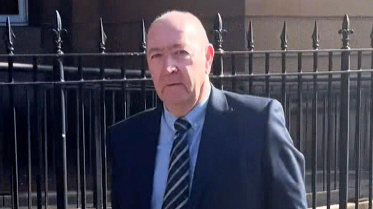 A balding man wearing a dark blue suit and a striped tie walking in front of a wrought iron fence