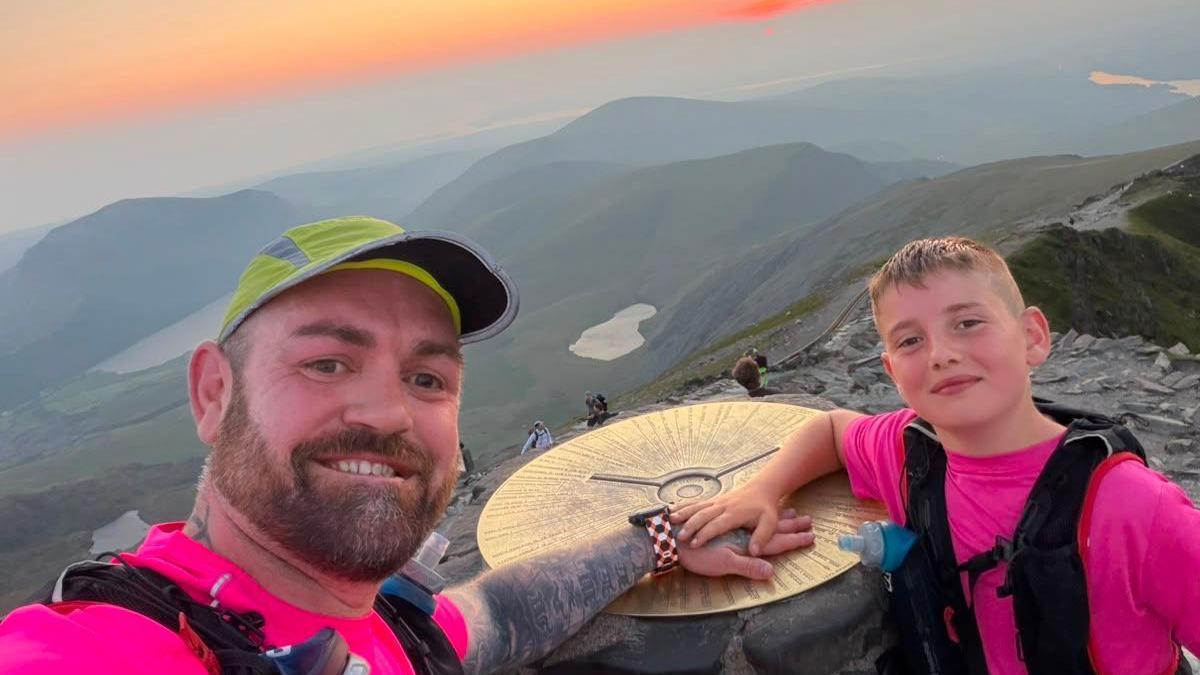 Daniel Thomas and Albie-Junior standing at the peak of Yr Wyddfa, also known as Snowdon. An organe and pink sunset can be seen in the horizon. They are both smiling and looking at the camera.