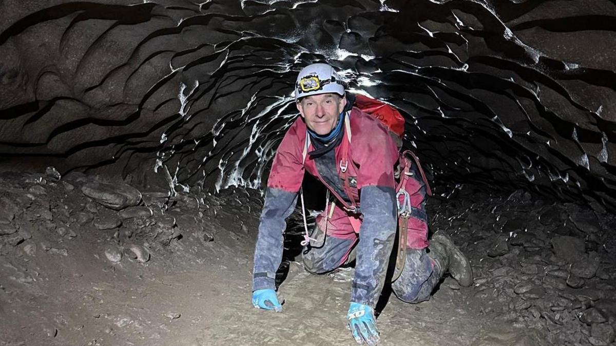 A man wearing protective outdoor clothing and a helmet with a torch on his hands and knees inside a tight, rocky space.