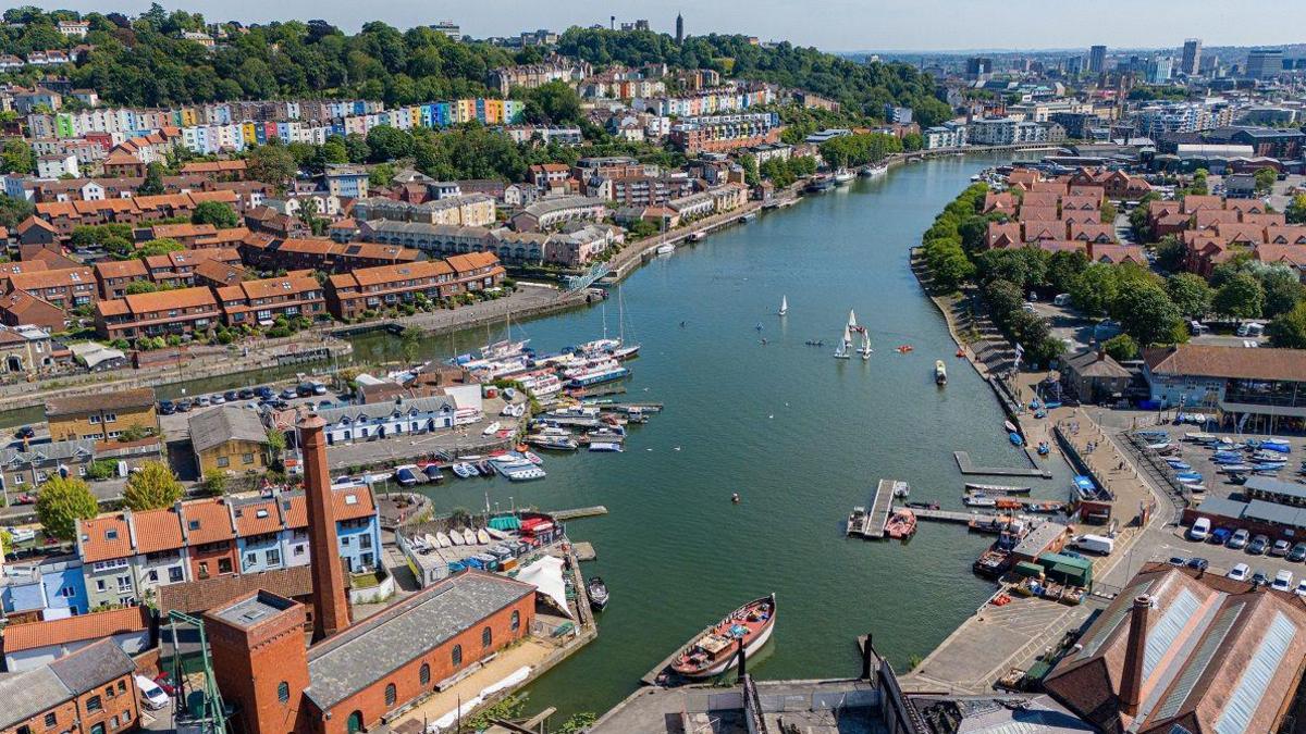 An aerial view of Bristol Harbour from a drone over Underfall Yard. Sailing dinghies are visible in the foreground and in the distance you can see the skyscrapers of the city centre