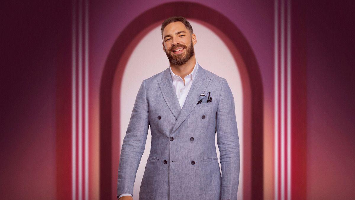 Publicity shot featuring Steven Smith in a grey jacket and white shirt smiling at the camera, framed by a pink archway