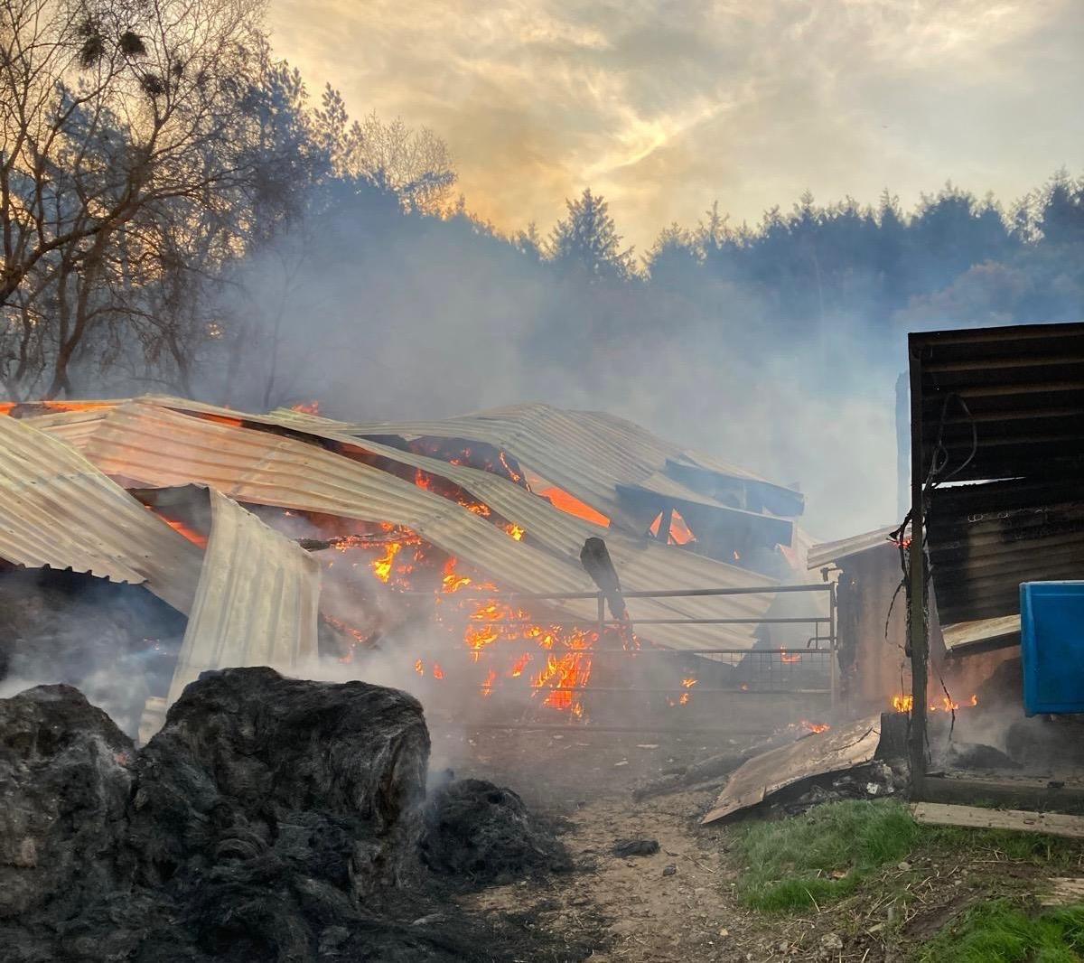 A metal outbuilding has completely collapsed. Bright orange flames can still be seen raging within.