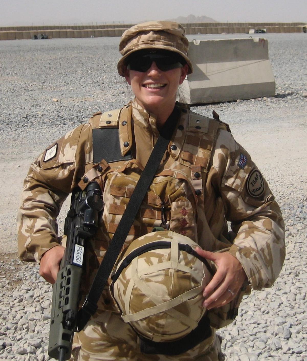 Bex is wearing combat gear and holding a rifle and helmet