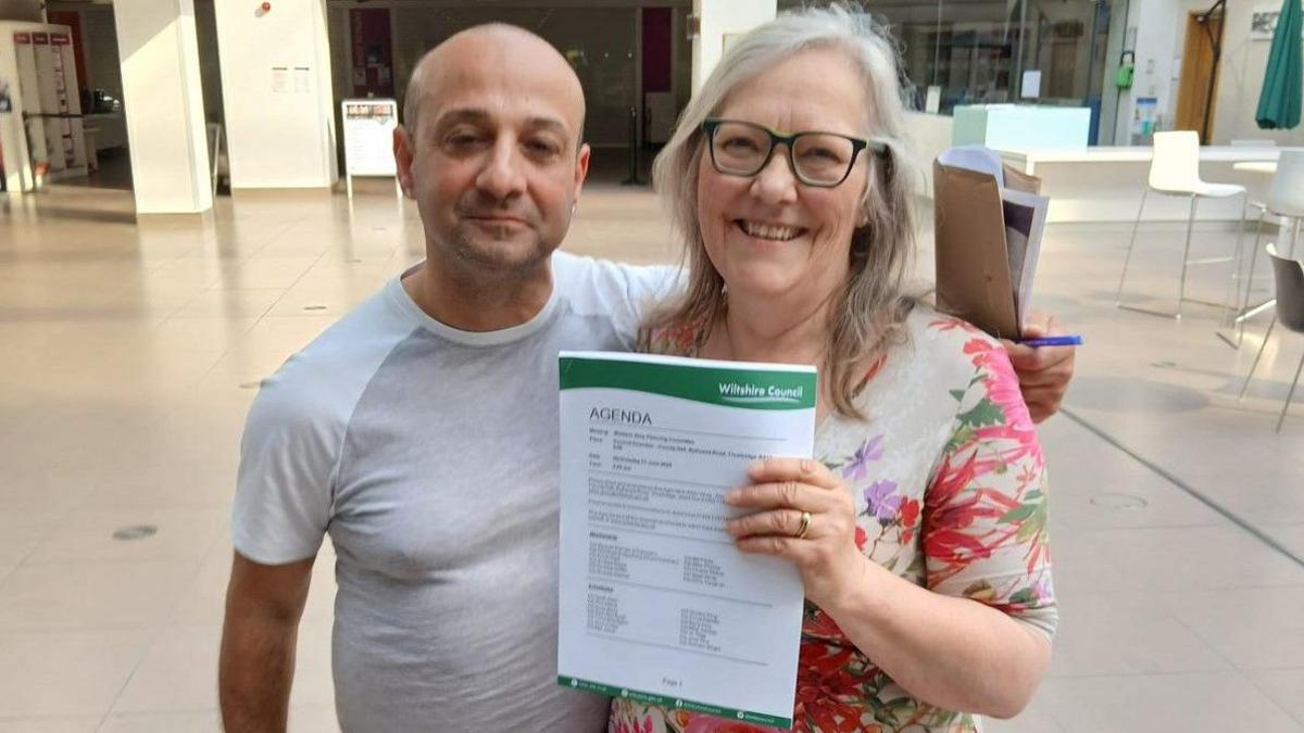 Kevin and Lizzie smile at the camera from the Wiltshire Council foyer area. They are holding Wiltshire Council branded paper to the camera showing the meeting agenda