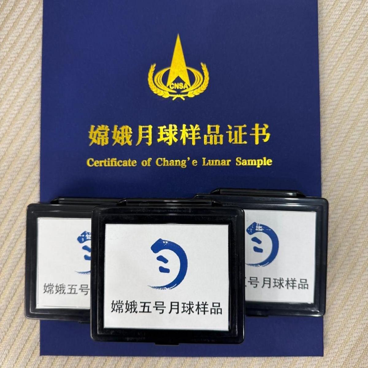 A blue folder that contains a certification from the Chinese space agency about the Chang'e 5 samples. On top are three boxes with white background and black borders. On the front is the Chinese space agency Chang'e logo and writing in Chinese script.