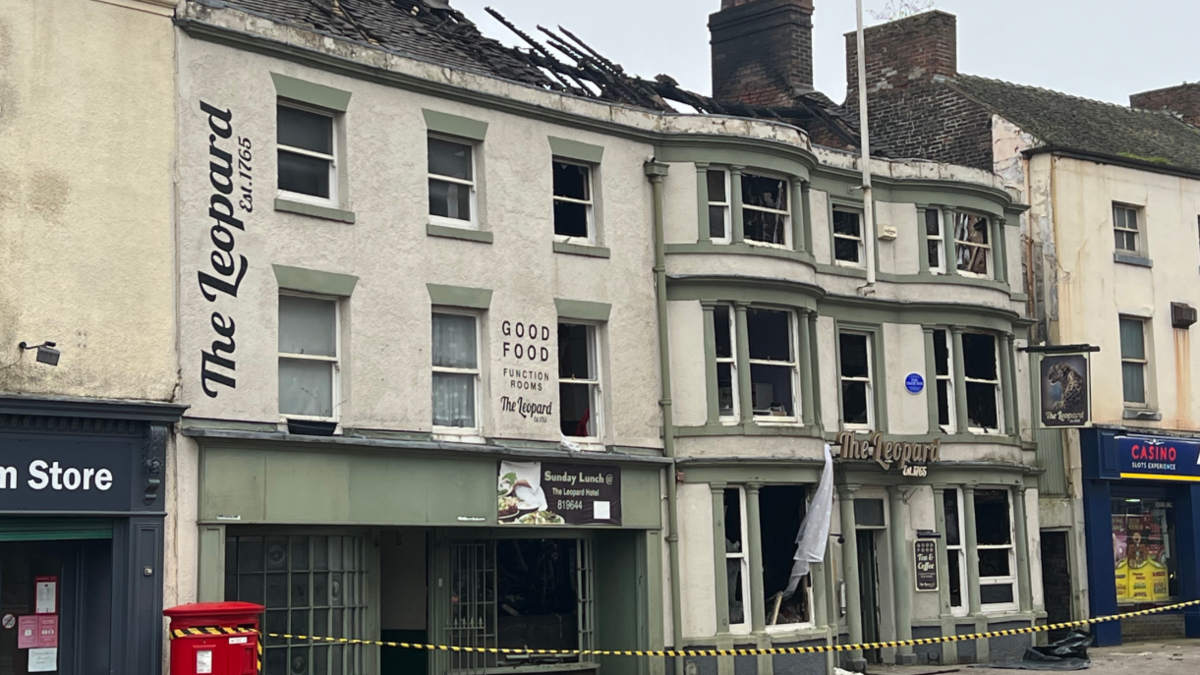 A white building with green trim, its roof apparently damaged, sits on a street between other shops. "The Leopard - est. 1765" is printed on the front, while other letting advertises food and function rooms. A black and yellow cordon 